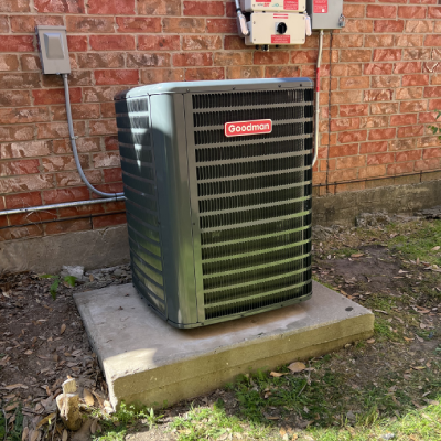 A Goodman air conditioning unit sits on a concrete slab beside a brick wall, with utility panels visible in the background A Goodman air conditioning unit sits on a concrete slab beside a brick wall, with utility panels visible in the background.